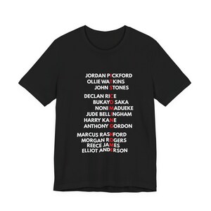 May include: A black short-sleeved t-shirt featuring a list of names printed in white text. The names include Jordan Pickford, Ollie Watkins, John Stones, Declan Rice, Bukayo Saka, Noni Madueke, Jude Bellingham, Harry Kane, Anthony Gordon, Marcus Rashford, Morgan Rogers, Reece James, and Elliot Anderson. The letters forming the phrase ITS COMING HOME are highlighted in red vertically down the centre of the list.