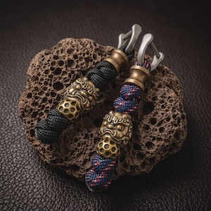Könnte beinhalten: Zwei Paracord-Anhänger mit metallischen Perlen in Form stilisierter Löwenköpfe und kugelförmigen Elementen mit kreisförmigen Aussparungen. Jeder Anhänger hat einen silberfarbenen Metallschäkel und besteht aus geflochtener Schnur in Schwarz und Blau-Rot-Weiß.