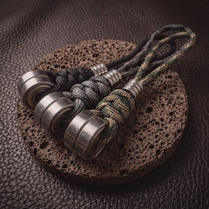 May include: Three zip pulls featuring silver-toned metal ball bearings attached to braided paracord loops. The cords are black, grey, and a camouflage pattern of green, tan, and black. Each pull includes a small, cylindrical metal bead connector.