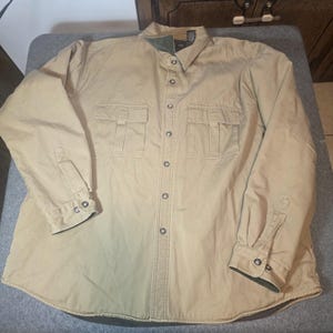 May include: A long-sleeve button-down shirt in a solid tan color. The shirt features a standard collar with a dark green inner lining, a full front button placket, and two chest pockets with buttoned flaps. The cuffs also have button closures.