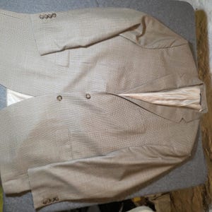 May include: A tan blazer featuring a fine brown micro-check pattern, a notched lapel, and a light beige interior lining. The garment includes two front buttons, two flap pockets, and four smaller buttons on each sleeve cuff for a structured look.