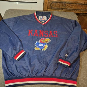 May include: A navy blue long-sleeve pullover windbreaker featuring the word KANSAS in red letters and a Jayhawk mascot graphic with the letters KU. The garment has a V-neck collar with red and white stripes, and matching striped cuffs and hem.