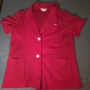 May include: A short-sleeved red blazer featuring a notched lapel, two white buttons for closure, two large patch pockets at the bottom, and a smaller patch pocket on the left chest. The interior neck label displays the text ARC Fashions.