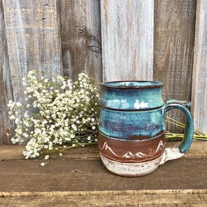 May include: A ceramic mug featuring a two-tone glaze with a glossy teal upper section and a matte brown lower section. The lower portion is decorated with a carved mountain range design. The mug has a sturdy handle and a wide, rounded base.