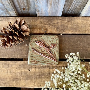 May include: A square ceramic wall plaque with a sage green glaze and a textured surface. The plaque features a raised, dark brown relief of a plant stem with a small bud and a tiny butterfly. A thin wire loop is attached to the top for hanging.