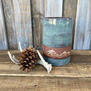 May include: A cylindrical ceramic mug featuring a speckled blue glaze on the top and bottom sections, separated by a central band of unglazed terracotta clay embossed with a detailed mountain range landscape, accompanied by a pinecone and antlers.