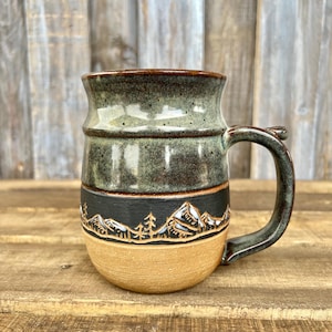 May include: A ceramic mug featuring a three-tone design with a speckled sage green top, a black middle band decorated with an etched mountain and pine tree landscape, and a solid tan base. The mug includes a curved handle on the right side.