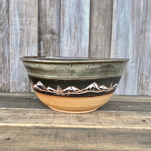 May include: A ceramic bowl featuring a wide rim with a speckled sage green glaze, a black middle band decorated with a carved mountain range and pine tree motif, and a natural tan unglazed base with a smooth, rounded bottom silhouette.