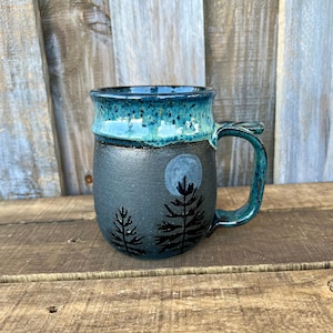 May include: A ceramic mug with a matte dark gray base featuring black painted pine trees and a pale gray moon. The upper portion of the mug and the handle are finished in a glossy, speckled teal glaze that drips down onto the dark gray body.