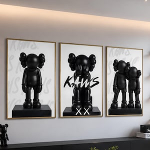 Peut inclure: Un ensemble de trois impressions d'art mural encadrées représentant des personnages KAWS noirs sur fond blanc. L'impression centrale affiche le texte KAWS et XX. L'œuvre est exposée sur un mur au-dessus d'un long meuble média noir.