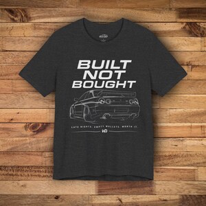 Könnte beinhalten: Ein anthrazitfarbenes Kurzarm-T-Shirt mit einer weißen Strichzeichnung der Rückansicht eines Sportwagens. Der Text BUILT NOT BOUGHT steht über dem Auto und LATE NIGHTS. EMPTY WALLETS. WORTH IT. darunter, ergänzt durch ein kleines WD-Logo.