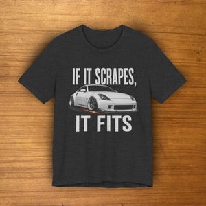 Puede incluir: Una camiseta de manga corta gris carbón oscuro con un gráfico de un coche deportivo blanco. El texto sobre el coche dice IF IT SCRAPES, y el texto debajo del coche dice IT FITS. El diseño está centrado en la parte delantera de la camiseta.