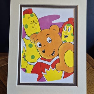 May include: A matted art print featuring a cartoon illustration of a brown bear wearing a red and yellow cape, positioned in front of two yellow characters with green polka dots, one with a red mohawk and the other with a red helmet-like hat.