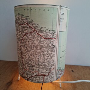 May include: A cylindrical lamp with a vintage map design. The lamp shade features a detailed map with place names and red lines. The lamp has a white base with three legs and a white power cord. The text "STOL CHANNEL" and "MINEHEAD" are visible.
