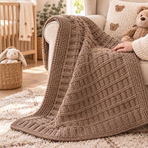 Può includere: Una coperta in maglia grossa di colore marrone medio tinta unita, caratterizzata da un motivo a griglia testurizzato con un ampio bordo a coste. La coperta è drappeggiata su un mobile, mettendo in risalto la trama del filato spesso.