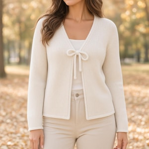 May include: A cream-colored long-sleeve knit cardigan featuring a V-neckline and a single tie closure at the center front. The garment is worn over a plain white top and paired with light-colored trousers, showcasing a textured knit pattern.