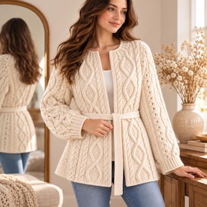 Könnte beinhalten: Eine cremeweiße Strickjacke mit Zopfmuster, rundem Halsausschnitt, langen Ärmeln mit gerippten Bündchen und einem passenden Bindegürtel in der Taille. Das Kleidungsstück ist mit einem strukturierten Muster aus vertikalen Zöpfen und Strickmaschen gefertigt.
