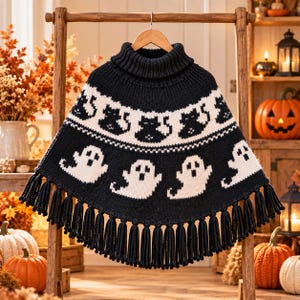 May include: A black knit poncho featuring a high turtleneck collar and a fringe hem. The garment displays a white horizontal band with a repeating black cat pattern, followed by a row of white ghost figures against the black knit background.