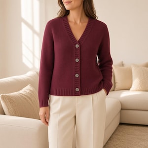 May include: A deep burgundy long-sleeve cardigan featuring a V-neckline, a ribbed knit trim along the front placket, cuffs, and hem, and four matching buttons down the center. The garment is paired with light cream-colored trousers.