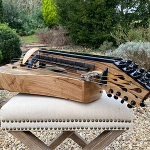 Op de afbeelding: Een houten nyckelharpa met een natuurlijke lichtbruine houtnerf, een zwarte toets met donkere toetsen en een zwarte stemkop met meerdere stempennen. Het snaarinstrument heeft een gebogen houten kam en metalen snaren over de klankkast.