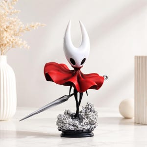 May include: A figurine of a character with a white, horned head and black eyes, wearing a flowing red cloak. The figure holds a long, silver, needle-like weapon and stands on a textured, grey, rocky base atop a black circular pedestal.