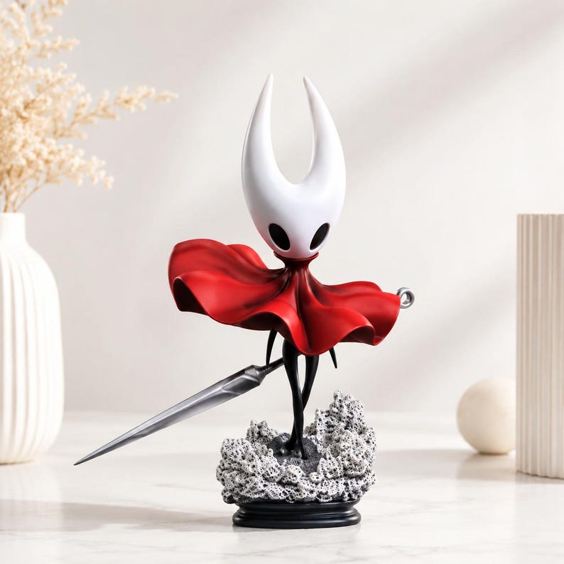 May include: A figurine of a character with a white, horned head and black eyes, wearing a flowing red cloak. The figure holds a long, silver, needle-like weapon and stands on a textured, grey, rocky base atop a black circular pedestal.