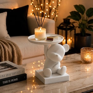 Peut inclure: Une table d'appoint en forme d'ours blanc composée d'une figurine d'ours aux formes arrondies assise sur une base carrée, soutenant un plateau circulaire au-dessus de sa tête. L'objet est fabriqué dans un matériau blanc mat et solide.