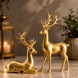 May include: Two gold-colored deer figurines with smooth, metallic finishes. One deer is depicted in a resting, lying-down position, while the other stands upright. Both figures feature detailed, branching antlers and are crafted with a solid, uniform gold tone.
