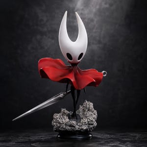 May include: A figurine of a character with a white, horned head and black oval eyes, wearing a flowing red cloak. The figure holds a long, slender silver needle-like sword and stands upon a textured, gray, rocky base against a dark background.