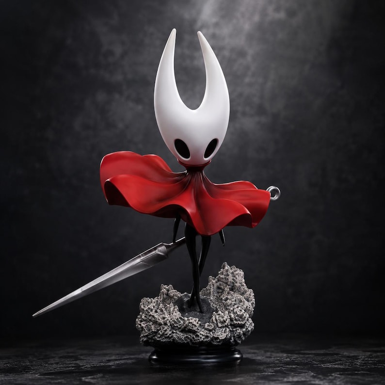 May include: A figurine of a character with a white, horned head and black oval eyes, wearing a flowing red cloak. The figure holds a long, slender silver needle-like sword and stands upon a textured, gray, rocky base against a dark background.