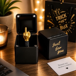 Puede incluir: Dos cajas de regalo cuadradas negras, una abierta y otra cerrada. La caja abierta contiene una figura de mano dorada con el dedo corazón extendido sobre un muelle metálico. La caja cerrada tiene el texto dorado Just for you en el lateral.
