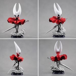 May include: A collectible figurine of a character with a white, horned head and black eyes, wearing a vibrant red cloak. The figure holds a long, silver-colored needle-like weapon and stands on a base decorated with sculpted gray roses.