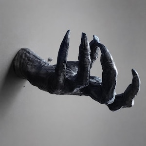 May include: A wall-mounted sculpture of a dark, textured creature hand with long, sharp, curved claws. The hand is positioned as if reaching forward, featuring detailed skin folds and a matte black finish that extends from the base to the fingertips.