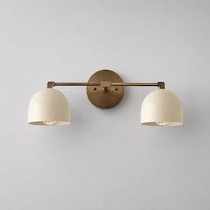 May include: A two-light wall sconce featuring a circular antique brass backplate and a horizontal bar supporting two dome-shaped off-white shades. The fixture has a metallic finish on the central arm and mounting plate with matte light-coloured covers.
