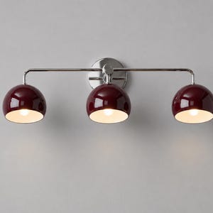 Pode incluir: Um candeeiro de parede de três luzes com uma barra horizontal em cromo polido e uma placa de montagem circular. Três abajures brilhantes em forma de cúpula, de cor bordeaux profundo, estão fixados à barra, cada um com uma lâmpada.