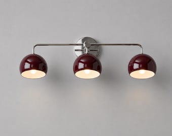 May include: A three-light wall fixture featuring a polished chrome horizontal bar and a circular mounting plate. Three glossy, deep burgundy dome-shaped shades are attached to the bar, each housing a single light bulb that emits a warm glow.