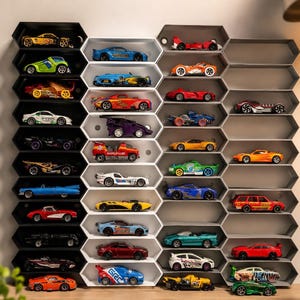 May include: A wall-mounted display unit composed of interlocking hexagonal compartments in black, white, and gray. The shelves hold a collection of various miniature toy cars in diverse colors, styles, and designs, arranged neatly within each individual cell.