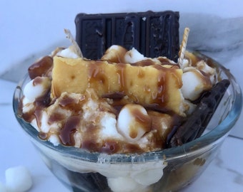 May include: A clear glass bowl filled with a dessert featuring a graham cracker base, topped with a square of golden-brown cake, marshmallows, chocolate, and drizzled with caramel sauce. Scattered around the bowl are graham crackers, chocolate bars, and marshmallows.