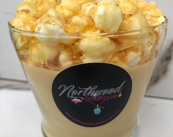 May include: A popcorn-themed candle in a clear glass. The candle is beige with a layer of caramel-colored wax and topped with popcorn. A black label with the text "Northwood Boutique" is on the glass.