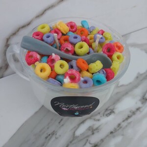 May include: A novelty candle shaped like a bowl of colorful cereal with a gray spoon. The candle is in a clear glass cup with a handle. The candle has a white base and is topped with colorful, ring-shaped wax pieces.