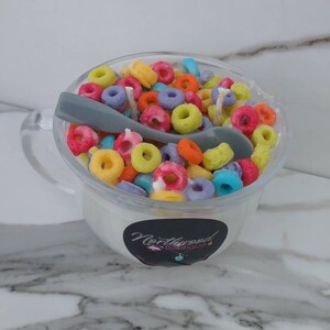 May include: A clear glass mug filled with a white substance, topped with colorful, ring-shaped cereal. A gray spoon rests inside. The mug has a handle and a black and white sticker that reads "Northwood Boutique."