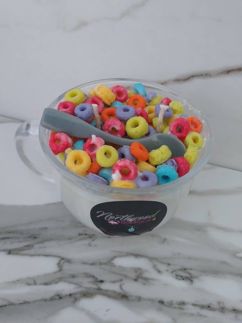 May include: A clear glass mug filled with a white substance, topped with colorful, ring-shaped cereal. A gray spoon rests inside. The mug has a handle and a black and white sticker that reads "Northwood Boutique."