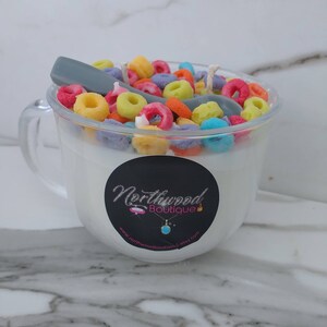 May include: A clear glass mug-shaped candle filled with white wax and colorful cereal pieces. A gray spoon rests on top. A black label with the text "Northwood Boutique" is on the front of the mug.