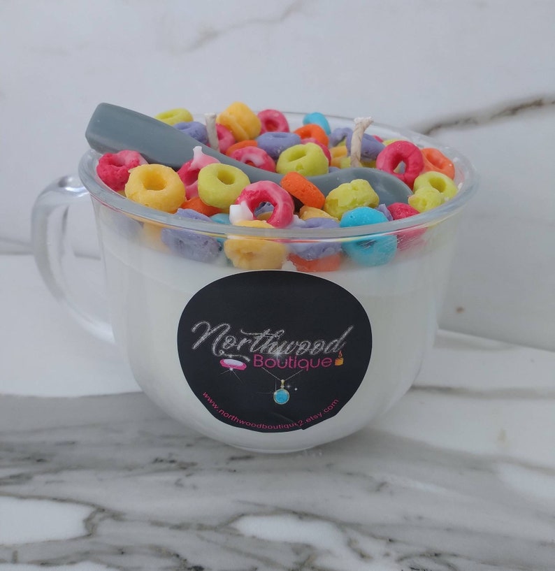 May include: A clear glass mug-shaped candle filled with white wax and colorful cereal pieces. A gray spoon rests on top. A black label with the text "Northwood Boutique" is on the front of the mug.