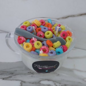 May include: A clear glass mug candle filled with colorful, cereal-shaped wax pieces. A gray spoon rests inside the mug. The candle has a white base and a black label with the text "Northwood Boutique".