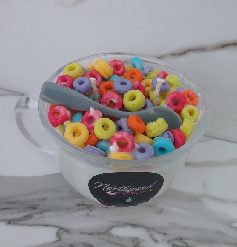 May include: A clear glass mug candle filled with colorful, cereal-shaped wax pieces. A gray spoon rests inside the mug. The candle has a white base and a black label with the text "Northwood Boutique".