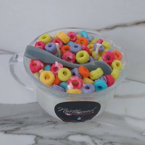 May include: A clear glass mug filled with a white substance and colorful, ring-shaped objects, resembling cereal. A gray spoon rests inside. The mug has a handle and a black and white sticker with the text "Northwood Boutique".