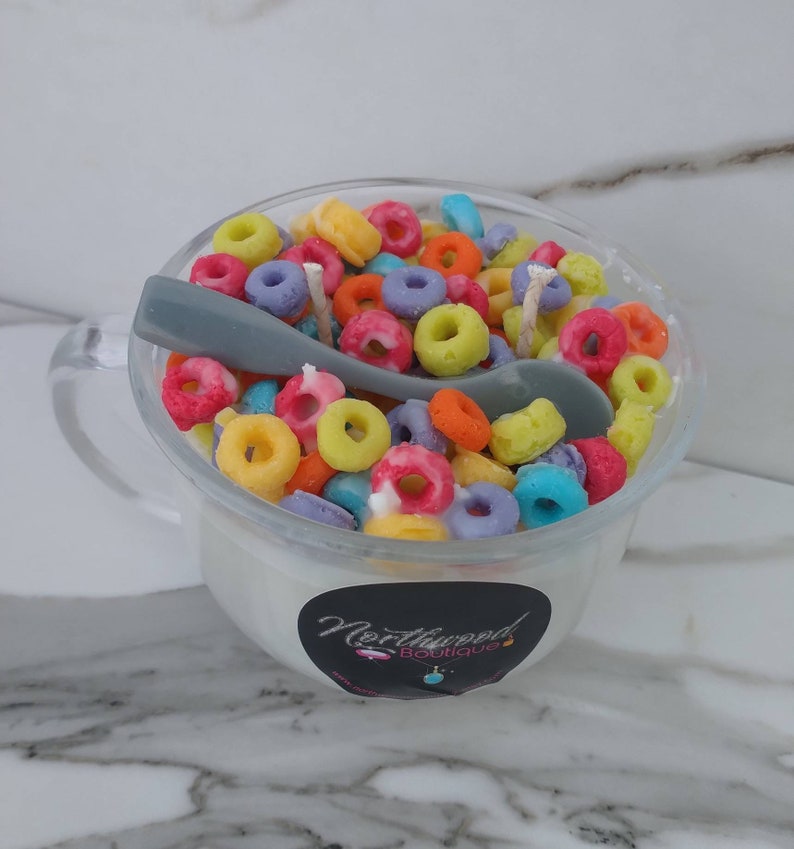 May include: A clear glass mug filled with a white substance and colorful, ring-shaped objects, resembling cereal. A gray spoon rests inside. The mug has a handle and a black and white sticker with the text "Northwood Boutique".