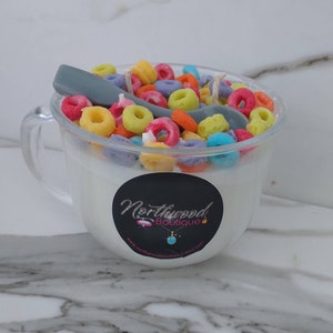 May include: A clear glass mug candle filled with white wax and colorful cereal pieces. A gray spoon rests on top. A black sticker with the text "Northwood Boutique" is on the front of the mug.