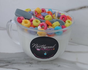 May include: A clear glass mug candle filled with white wax and colorful cereal pieces. A gray spoon rests on top. A black sticker with the text "Northwood Boutique" is on the front of the mug.